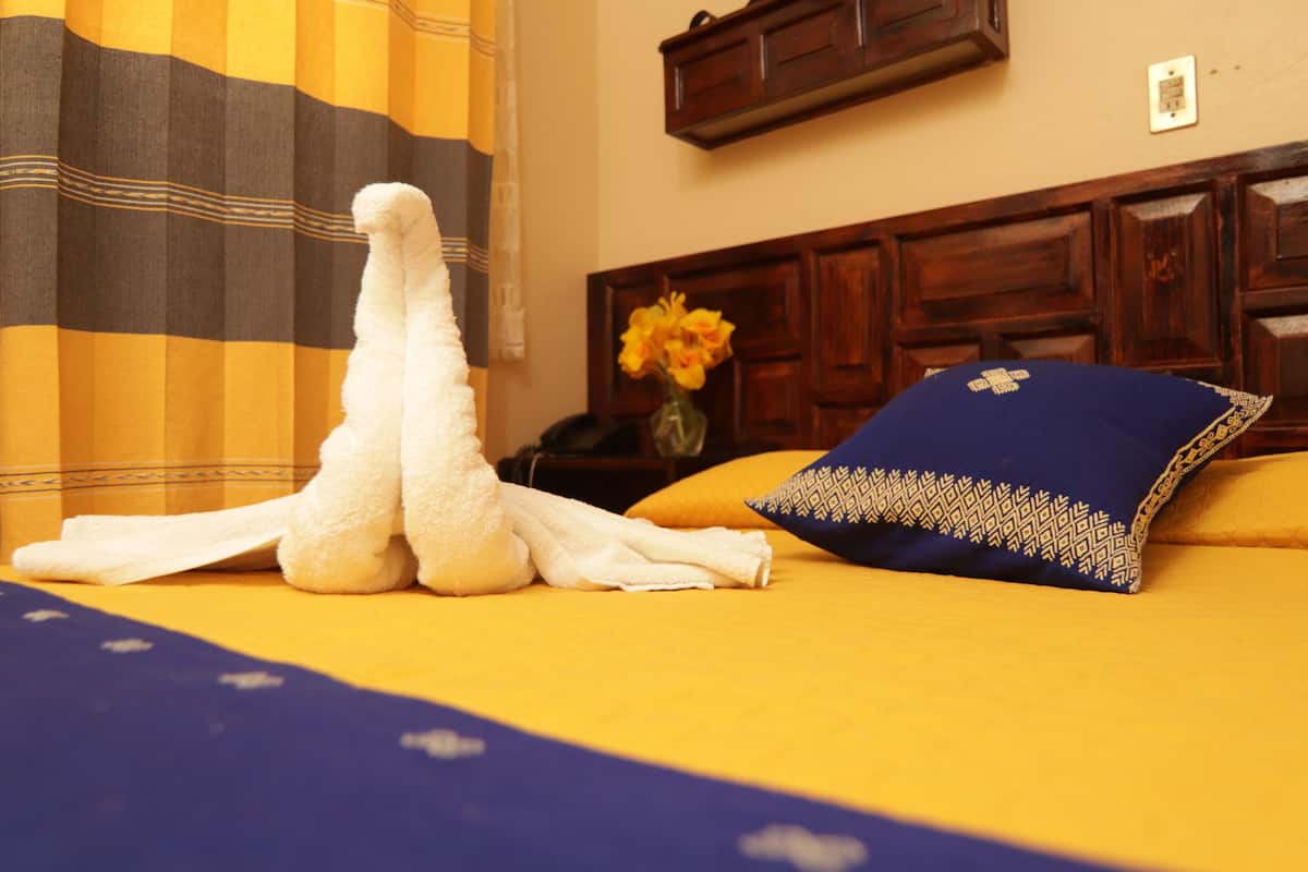 Ein Bett mit einer blau-gelben Tagesdecke, ein weißes Handtuch, ein blaues Kopfkissen mit gemustertem Rand und eine Vase mit gelben Blumen auf einem Nachttisch.