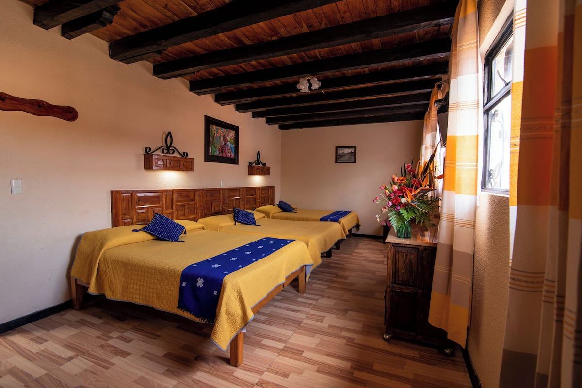 Ein Hotelzimmer mit Holzböden, einer hohen Decke mit sichtbaren Balken und einem Bett mit gelber Tagesdecke und blauem Kopfkissen.