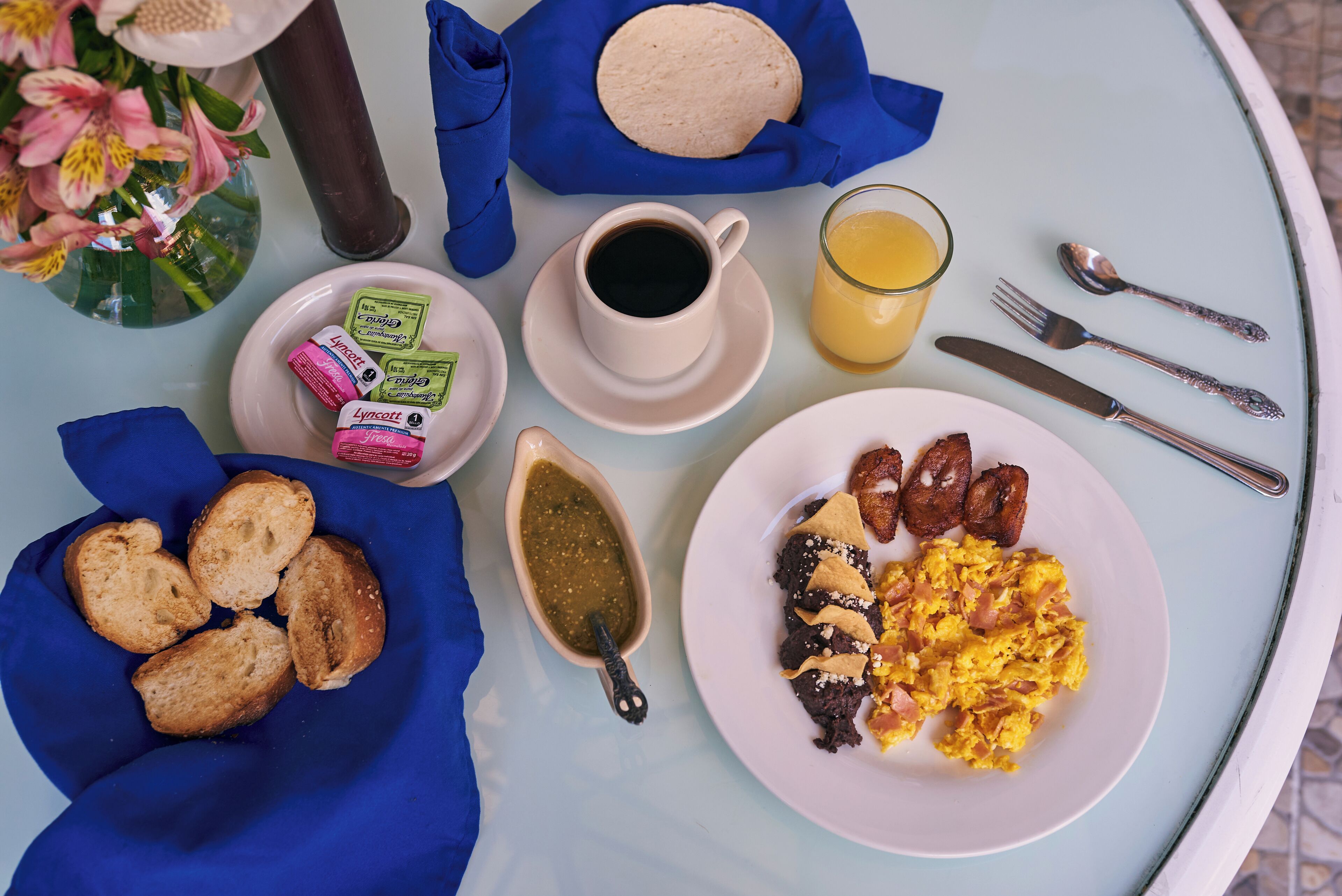 A breakfast table with a plate of scrambled eggs, refried beans, and tortillas, a cup of coffee, a glass of orange juice, and a bowl of salsa.