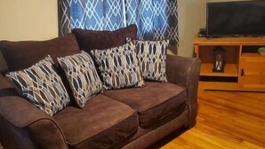 Un sofá marrón con cojines estampados, un televisor sobre un mueble de madera y una ventana con cortinas decoradas.