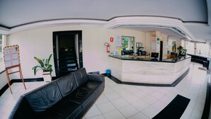 A modern hotel lobby with a reception desk, a black leather sofa, a small plant, and a fire extinguisher. - Diroma Internacional Resort - BVTUR (Caldas Novas)