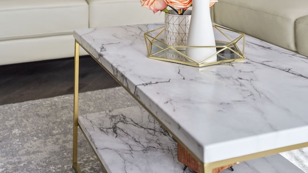 A living room with a marble coffee table, a white vase with flowers, a gold geometric tray, and a beige sofa.
