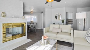 A modern living room with a white brick fireplace, a ceiling fan, a white sofa, and a dining area in the background.