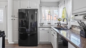 A modern kitchen with white cabinetry, a black refrigerator, a granite countertop, and a window with sheer curtains.