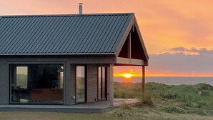 A modern house with a metal roof and large windows, overlooking a beach at sunset.