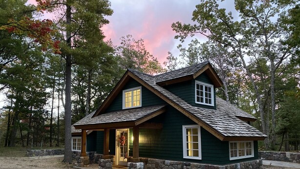 A two-story log cabin with a stone foundation, a wrap-around porch, and a stone wall surrounding the property. - The Little Green Cottage in the Woods (Glen Arbor)