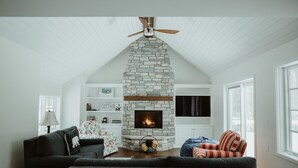 A cozy living room with a stone fireplace, a ceiling fan, and a large window. - The Little Green Cottage in the Woods (Glen Arbor)