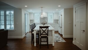 A modern kitchen with white cabinetry, a central island, and a dining area with dark chairs. - The Little Green Cottage in the Woods (Glen Arbor)