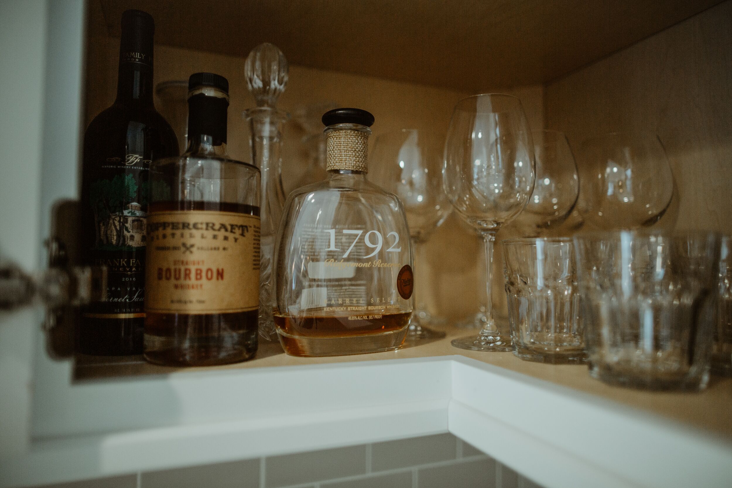 Une étagère contenant diverses bouteilles et différents types de verres, notamment une bouteille de bourbon et une bouteille portant l’étiquette « 1792 ».