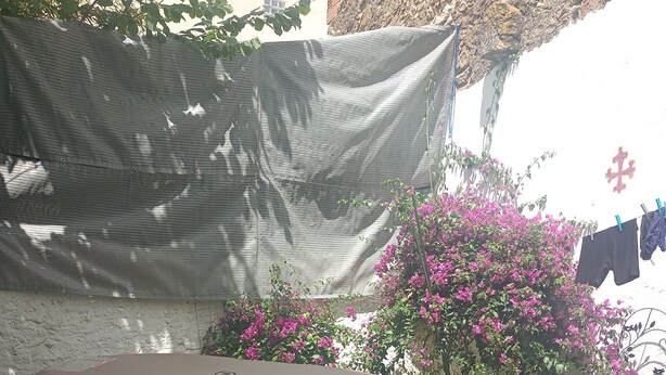 Un área de estar exterior en tonos beige con una mesa y sillas, rodeada por un jardín con flores rosadas y una planta trepadora.