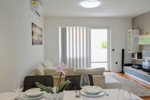 Een moderne woonkamer met een eethoek, een bank, een televisie en een groot raam met zonwering.