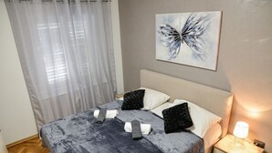 Ein ordentlich bezogenes Bett mit einer grauen Bettdecke, weißen Kissen und einem schwarzen Kissen. Ein Nachttisch mit einer Lampe und einem an der Wand hängenden Kunstwerk sind ebenfalls vorhanden.
