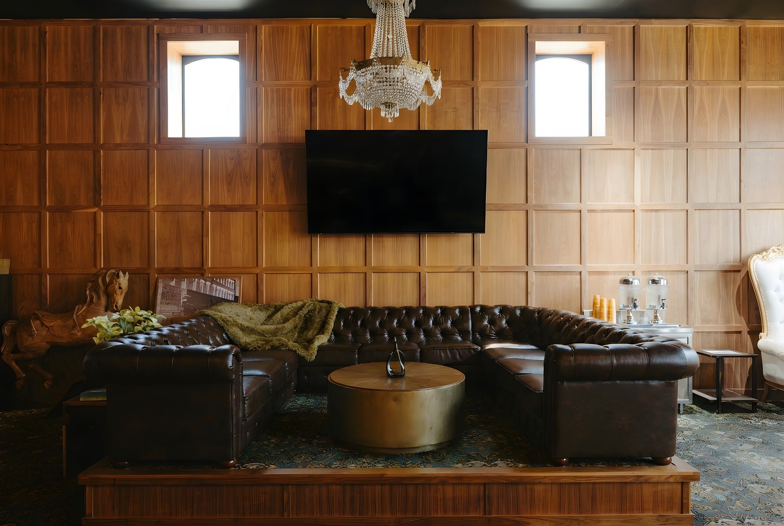 A living room with a leather sectional sofa, a round coffee table, a wooden accent wall with a flat-screen TV, and a chandelier.