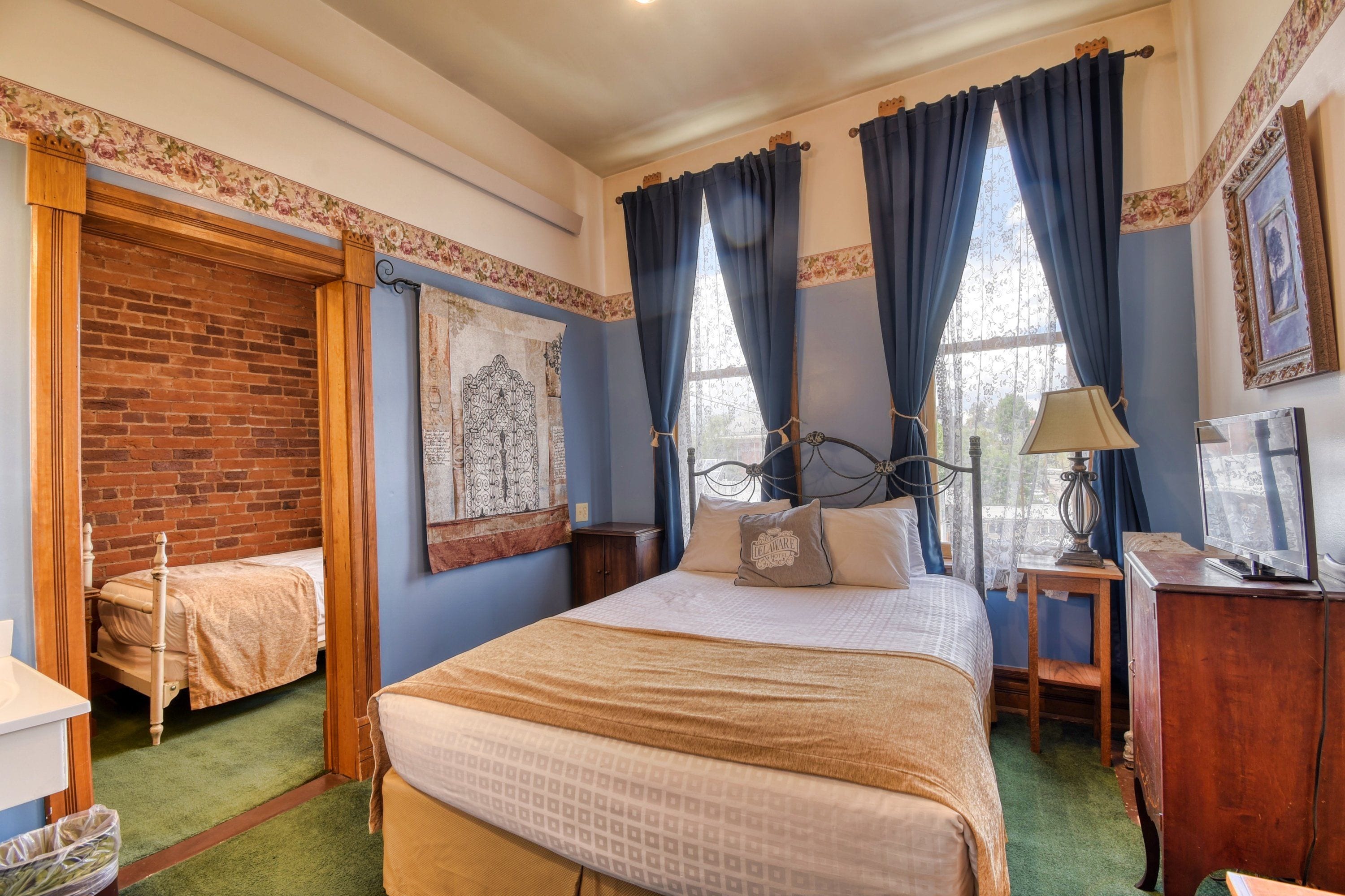 A hotel room with a large bed, a smaller bed, a brick wall, a window with curtains, a lamp, and a picture frame.