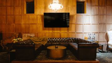 A room with a large flat-screen TV mounted on a wooden paneled wall, a leather sofa set, a round coffee table, and a chandelier.