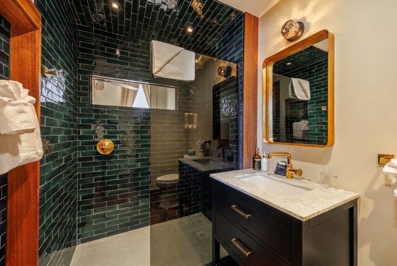 A bathroom with a dark green tiled wall, a white sink, a mirror, and a wooden door.