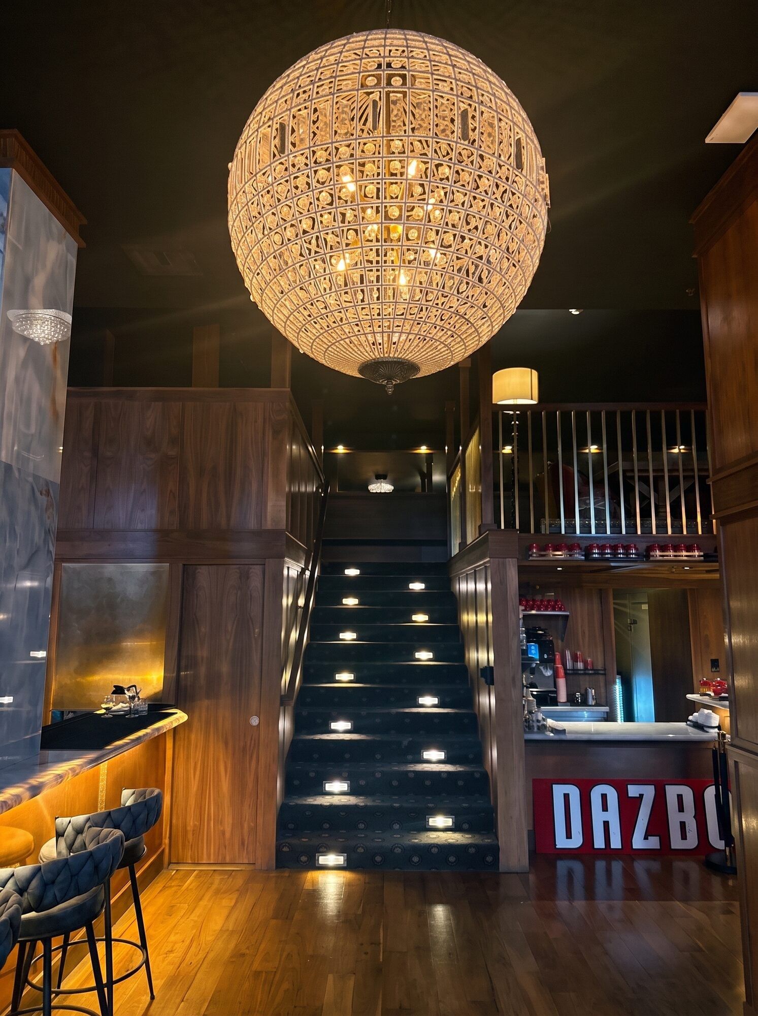 Una escalera con un candelabro, paredes de madera y una zona de bar con un letrero que dice 'DRAZBU'.