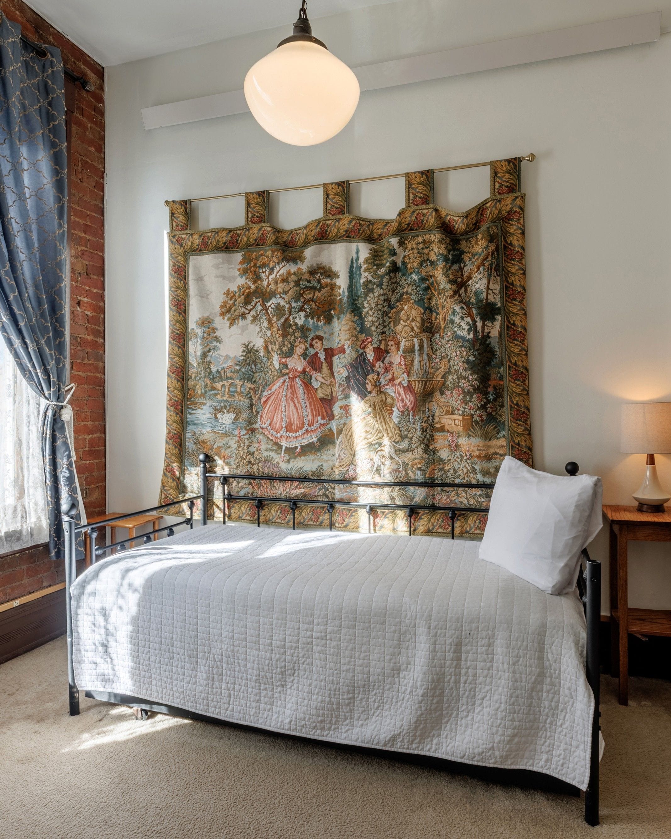 A bedroom with a bed, a tapestry, a lamp, and a window with curtains.