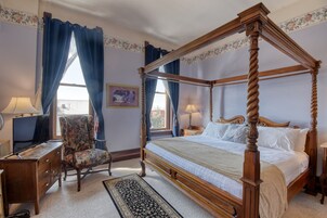 Uma cama com dossel e estrutura de madeira, uma poltrona com padrão floral, uma escrivaninha com luminária e um tapete no chão.