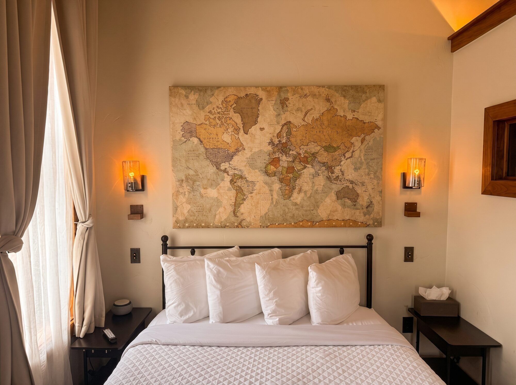 A bedroom with a large world map on the wall, a bed with white bedding, two bedside tables with lamps, and a window with curtains.