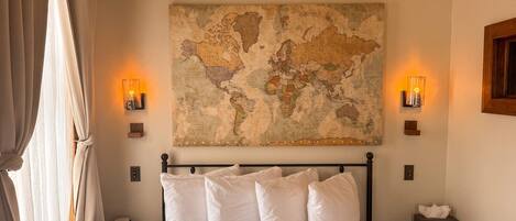 A bedroom with a large world map on the wall, a bed with white bedding, two bedside tables with lamps, and a window with curtains.