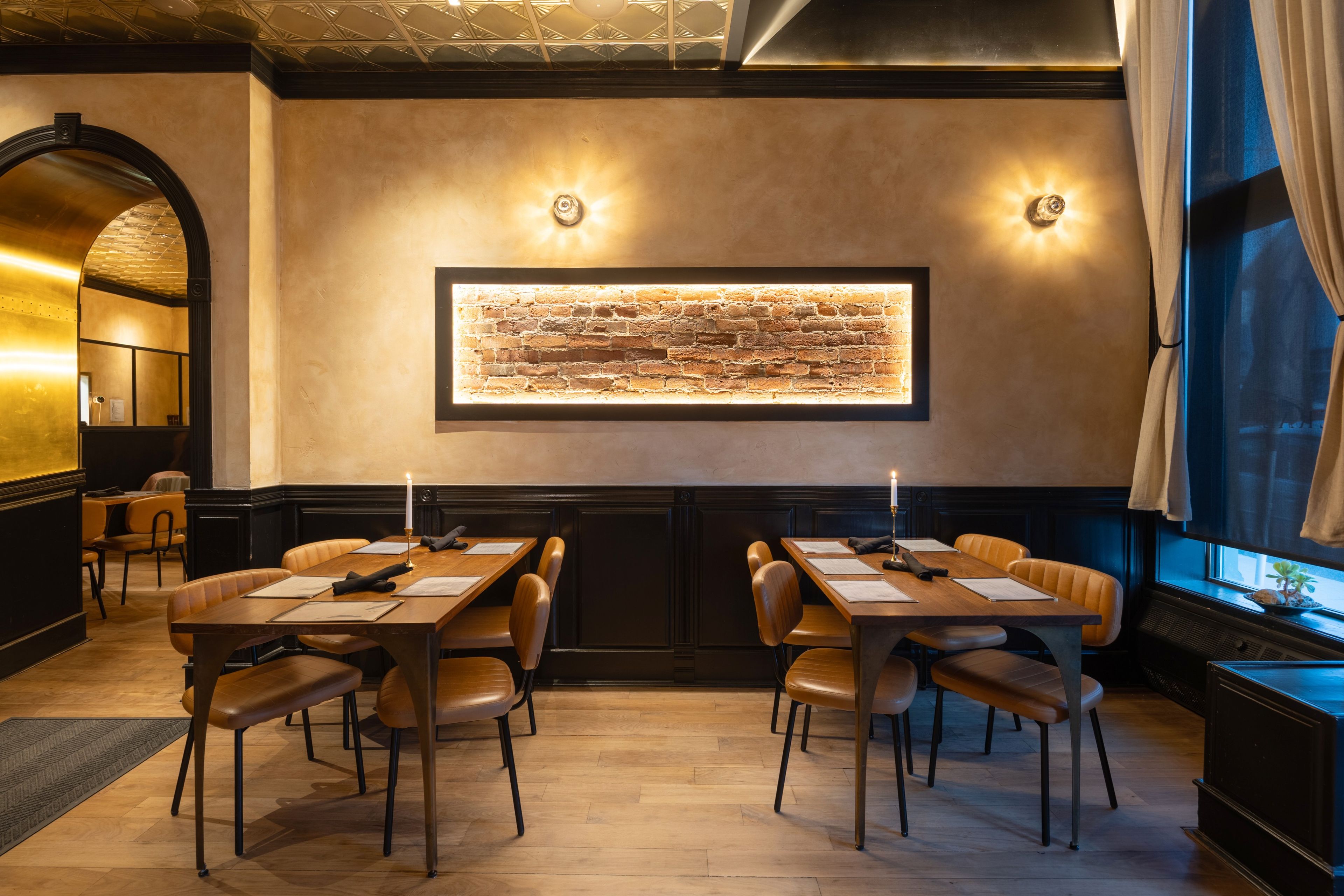 A restaurant with a long table and chairs, a brick wall with a framed picture, and a window with curtains.