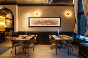 A restaurant with a long table and chairs, a brick wall with a framed picture, and a window with curtains.