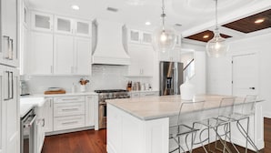 A modern kitchen with white cabinetry, a central island with bar stools, stainless steel appliances, and a wooden ceiling.