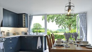A modern kitchen with dark cabinets, a large island, and a dining area with a view of greenery. - 2 bedroom accommodation in Beacon Cottage (Beacon Cottage)