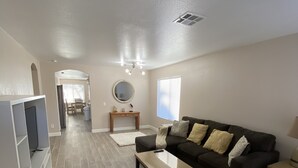A spacious living room with a dark brown sofa, a coffee table, and a TV stand. A round mirror hangs on the wall, and a window with blinds is visible. - One story, 3bedrooms. 15 min from Strip. Quiet & and safe. (Las Vegas)
