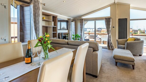 A spacious living area with a sofa, chairs, a dining table with a bottle and glasses, and a view of a water body through large windows. - 2 bedroom accommodation in Addlethorpe, near Skegness (Addlethorpe, near Skegness)