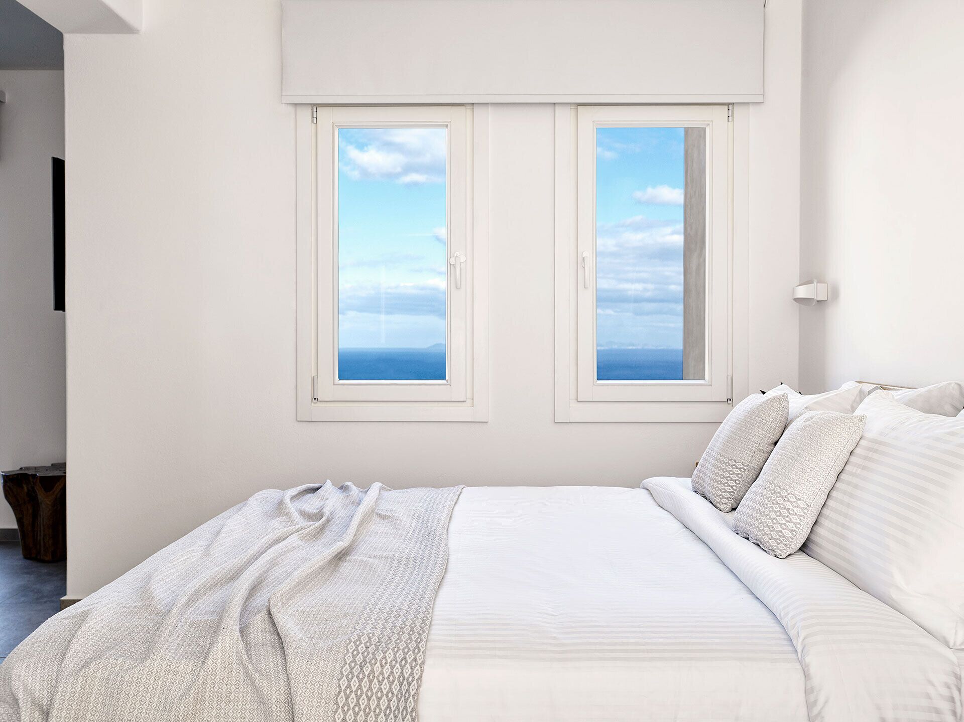 A bedroom with a large bed, two windows with a view of the sea, and a bedside table.
