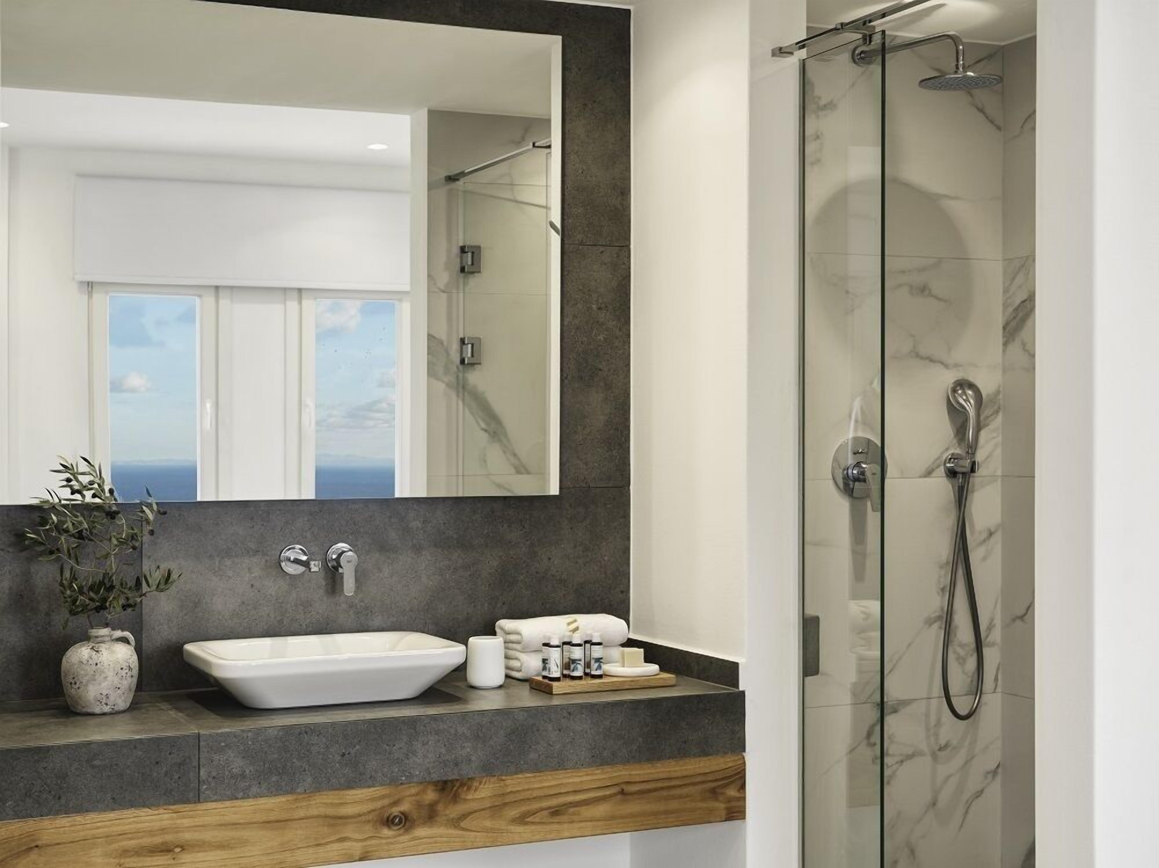 A modern bathroom with a large mirror, a white sink, a glass shower enclosure, and a view of the sea through the window.