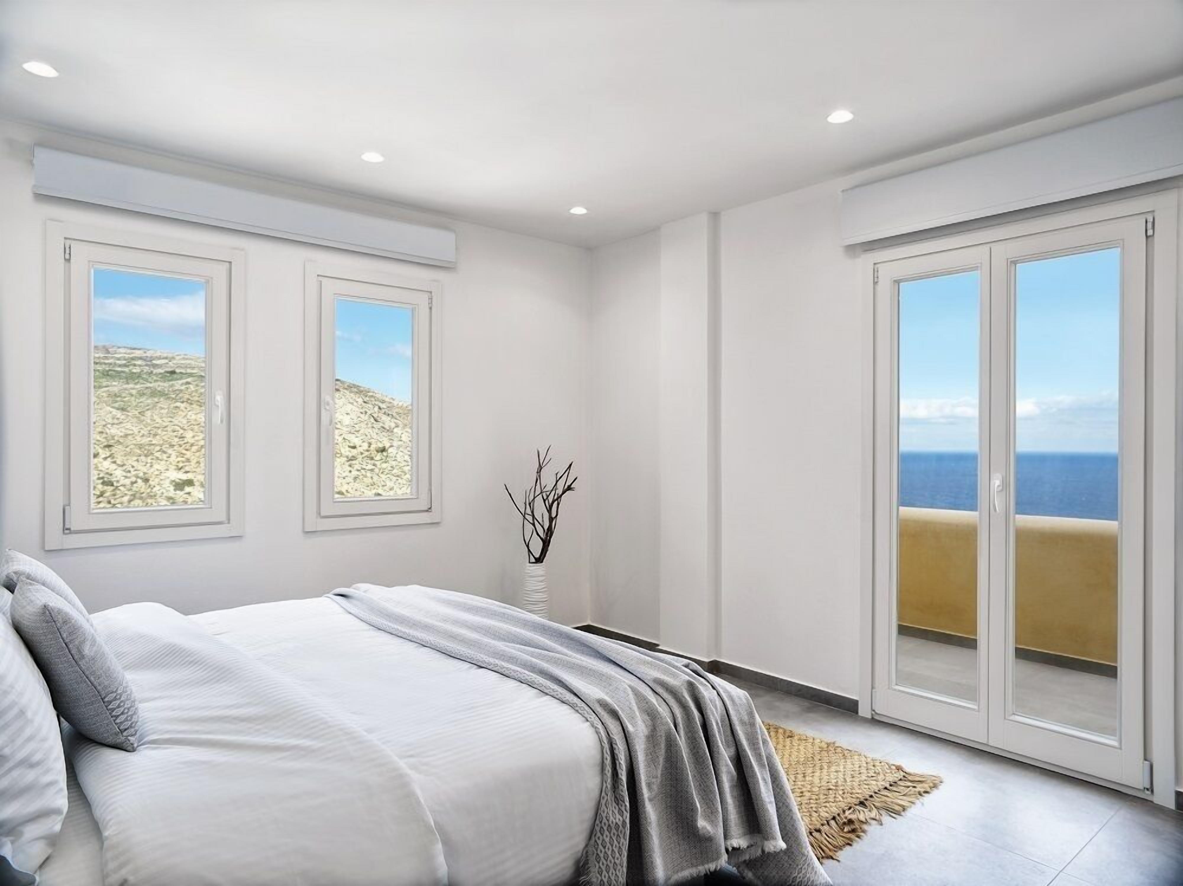 A bedroom with a bed, two windows with views of a rocky landscape, a vase with branches, and a sliding door to a balcony with a view of the sea.