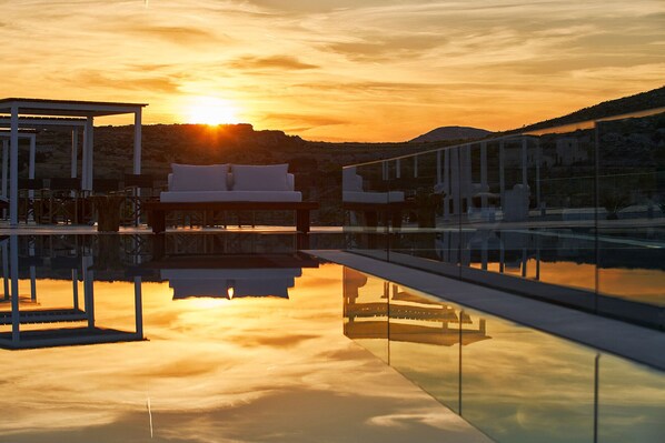 A serene pool area with a sunlit sky, a lounge with a sofa and chairs, and a clear reflection on the water. - AVATON Folegandros (Folegandros)