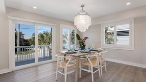 Un coin repas avec une table et des chaises en bois, un lustre et de grandes fenêtres équipées de stores.
