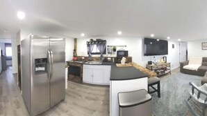 A modern kitchen with a refrigerator, sink, and dining area. There is a TV mounted on the wall and a sofa in the living area.