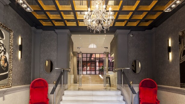 Uma escadaria imponente com um candelabro, cadeiras vermelhas e vista para uma biblioteca ao fundo.