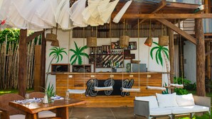 An outdoor bar area with wooden furniture, hanging white fabrics, and palm plants. - Amāra Beach Eco Hotel (Dibulla)