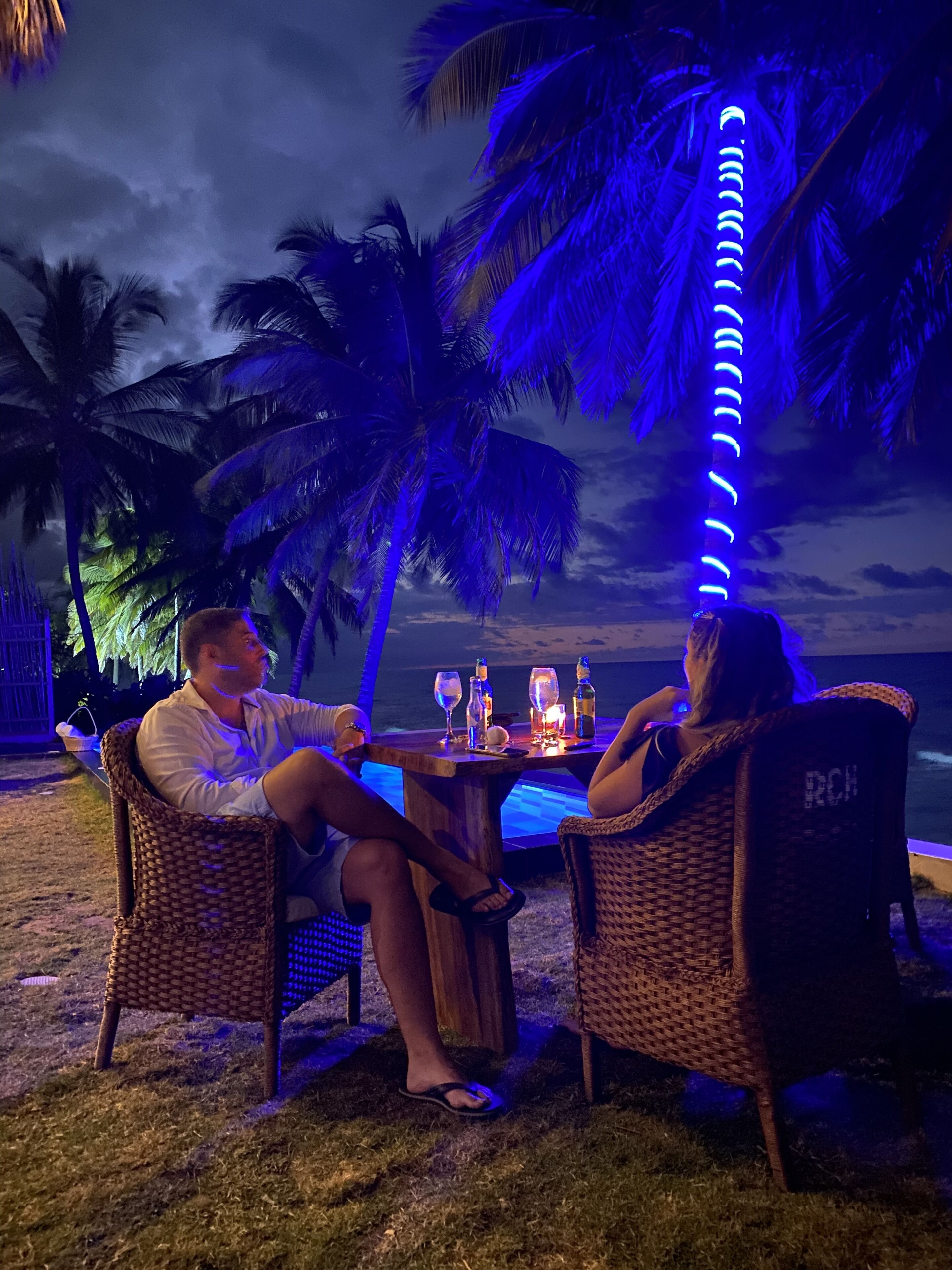 Two people seated at a wooden table with drinks, overlooking a beach with palm trees and a view of the ocean.