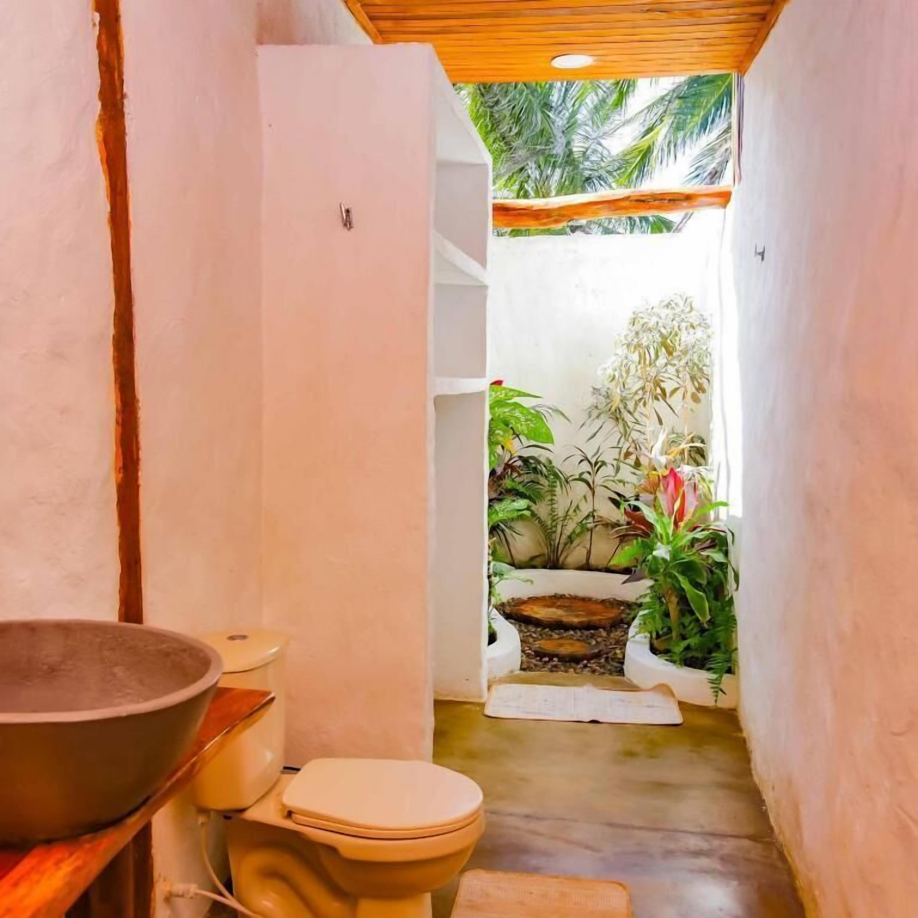 Une petite salle de bain comprenant des toilettes, un grand lavabo et un petit évier. Un petit jardin, avec des plantes et un élément décoratif aquatique, est visible à travers une porte ouverte.
