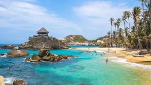 A beach with clear turquoise water, rocky outcrops, and a thatched-roof structure on a hill. - Amāra Beach Eco Hotel (Dibulla)
