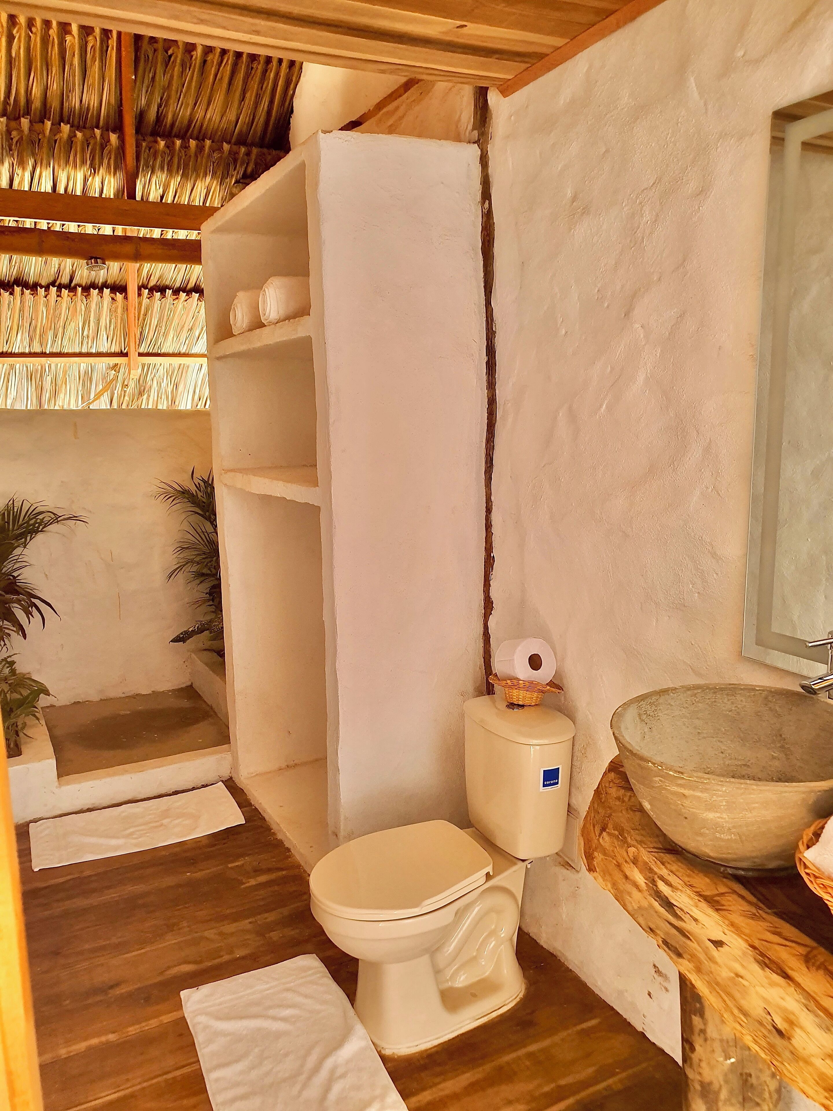 Ein Badezimmer mit Holzboden, Toilette in Weiß, einem großen Waschbecken aus Holz, einer Dusche mit weißem Duschvorhang und Ablage für Pflegeprodukte.