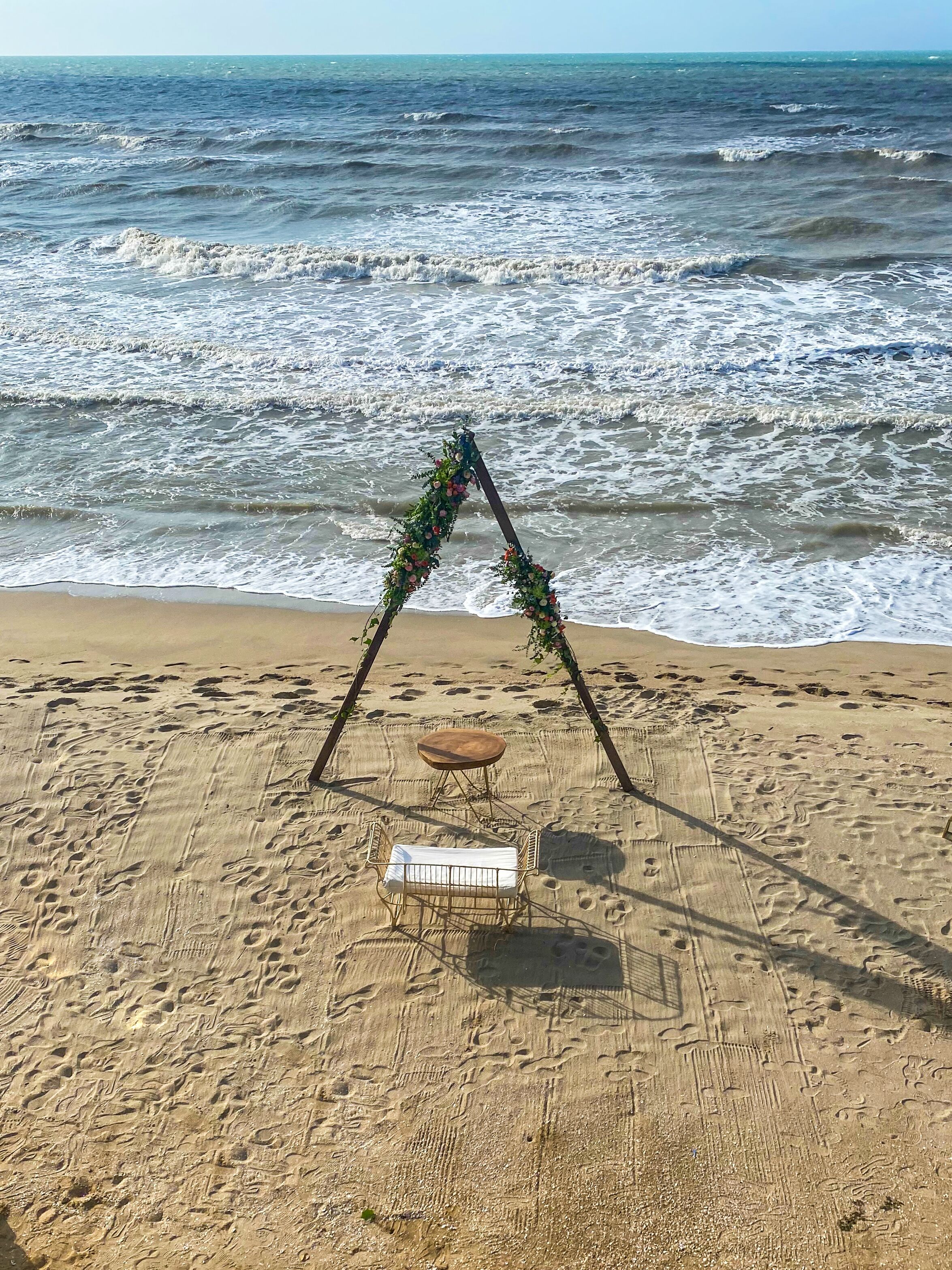 A beach with a wooden arch adorned with flowers, a small round table, and a metal chair.