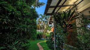 A pathway leading through a lush garden with various plants and a brick building in the background.