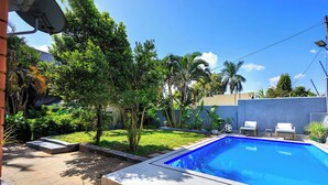 A swimming pool with a tiled deck, surrounded by greenery and a clear sky.