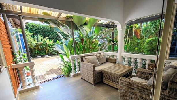 A covered patio with wicker furniture, a view of lush greenery, and a brick wall.