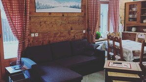 Tunnelmallinen mökin sisustus, jossa on puuseinä, sohva, ruokapöytä tuoleineen ja maalaus lumisesta maisemasta.