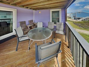 A covered outdoor seating area with a glass-top table and chairs, wooden deck flooring, and a view of a building with a blue exterior.
