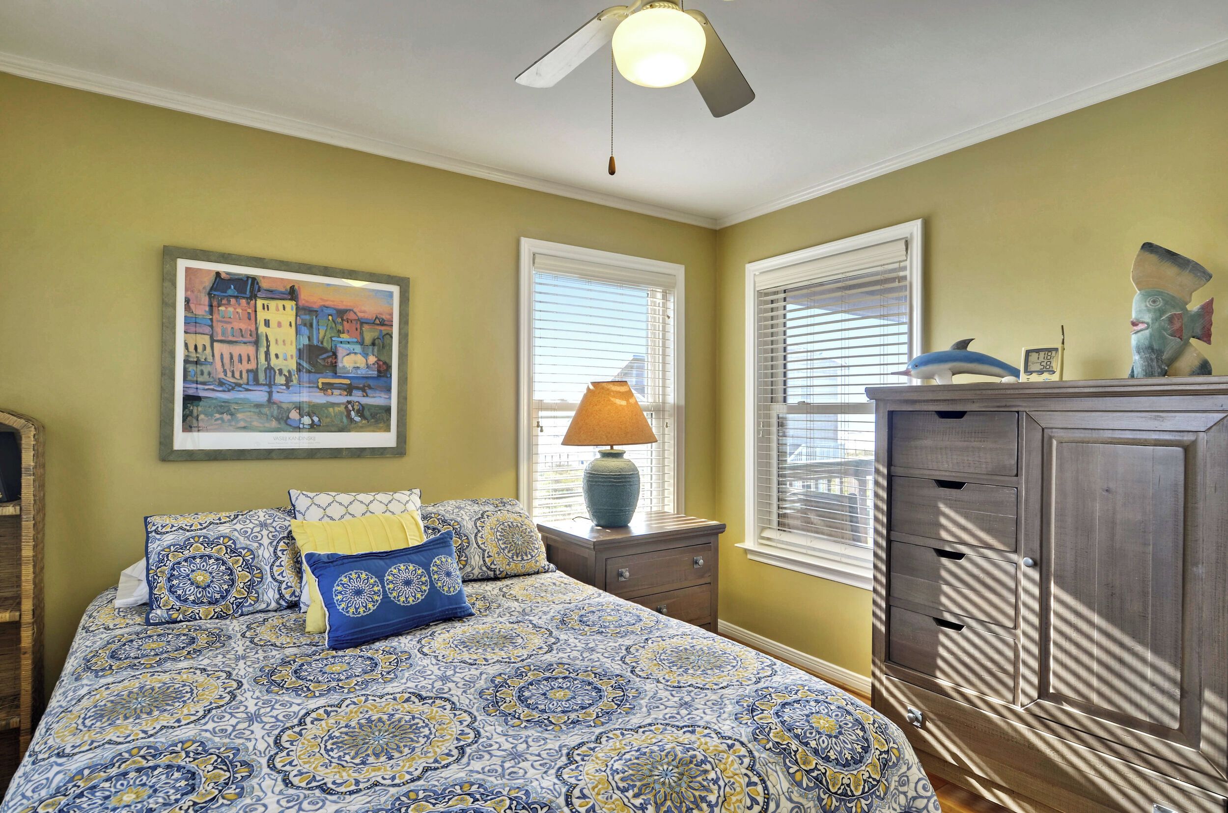 A bedroom with a patterned bedspread, a nightstand with a lamp, a dresser, and a painting on the wall.